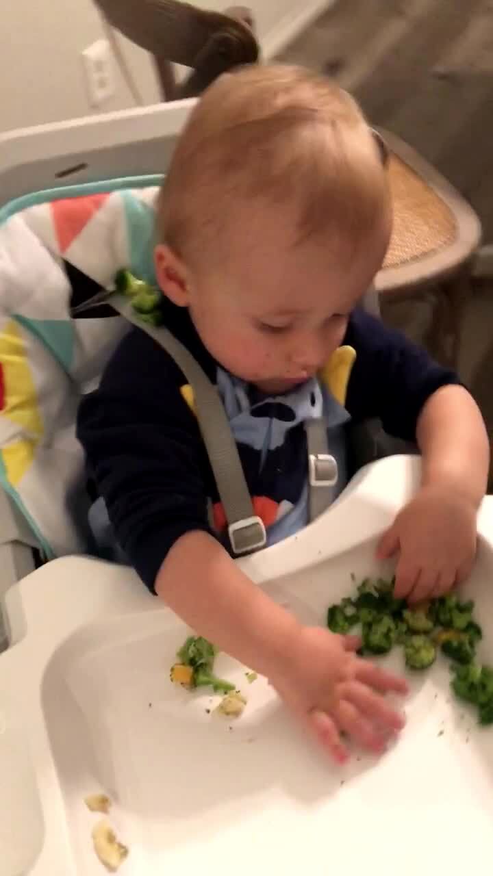 Video thumbnail showing: A tot boy hides broccoli behind his head while he sits in a high chair in the kitchen.
