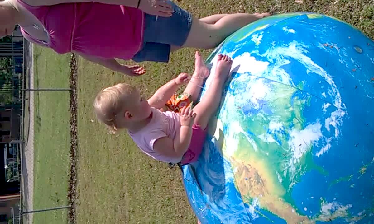 Video thumbnail showing: A tot girl sits on top of a large inflatable globe that fills with water in the yard. Her older sister leans against it and it pops and she falls into the water.