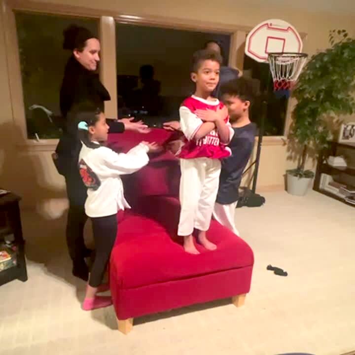 Video thumbnail showing: A young boy does a trust fall and falls forwards as the family reaches forward in the living room.